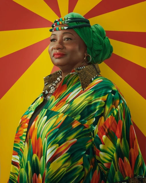 Portrait of a smiling mature Black woman with a green headwrap and colorful shirt against a backdrop with red and yellow rays.